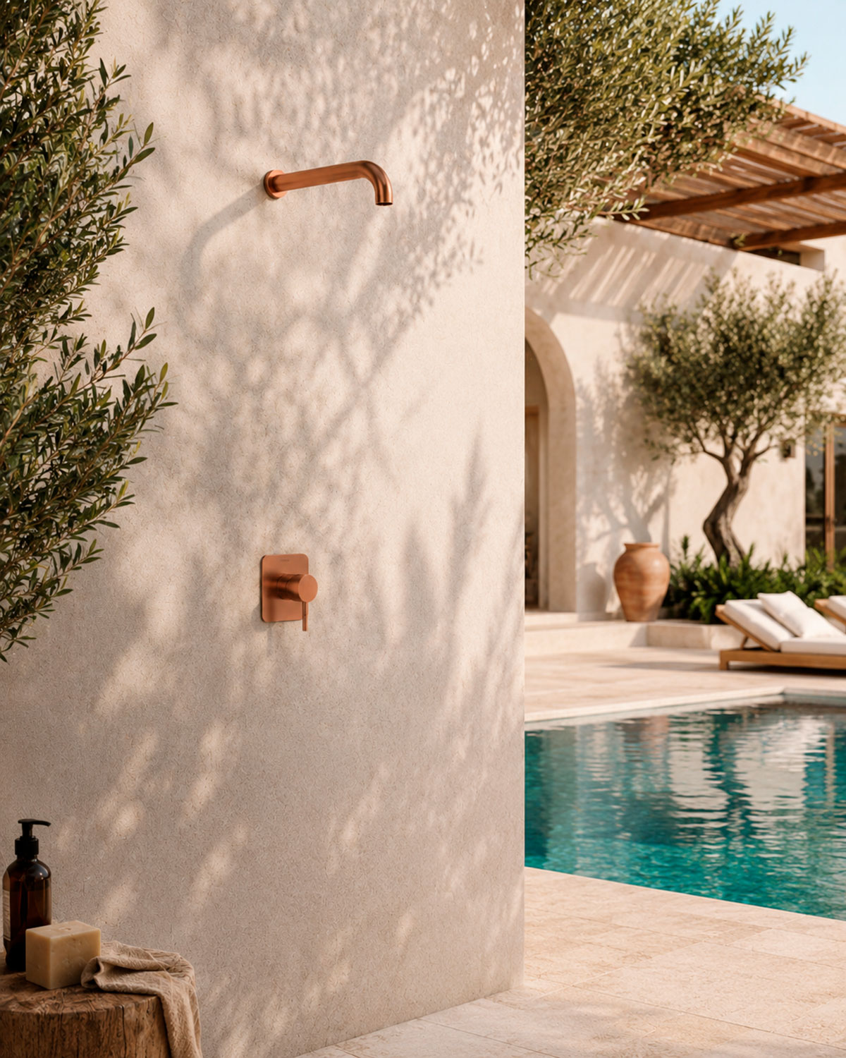 Ducha exterior de cobre Ramon Soler instalada entre un patio y una piscina con luz natural y vegetación mediterránea
